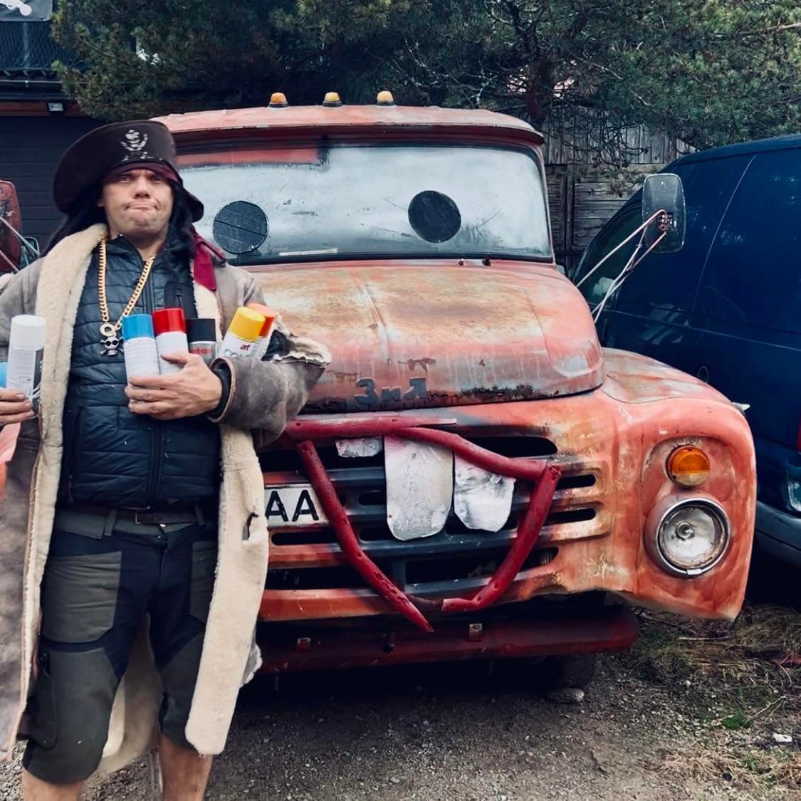 Pirate's Eye in a tricorn hat holding spray cans in front of a rusted orange ZIL truck with eyes painted on the windscreen